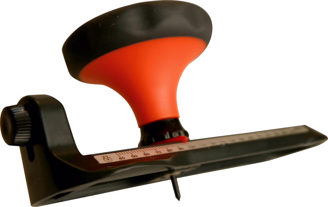 WOLFF_Dummy_15624_circular cutter_2025-08 Gros plan d'un outil à main pour le travail du bois (possiblement une ponceuse ou un rabot) doté d'une poignée ergonomique orange vif et d'un capuchon noir. Un bouton de réglage en plastique noir est visible sur le côté, et une règle métallique avec des chiffres et des graduations gravés longe la base. Une vis pointue ou une lame dépasse du dessous. L'outil est isolé sur un fond blanc, avec une faible profondeur de champ qui met en valeur la poignée et la règle, tandis que les autres parties sont légèrement floues.