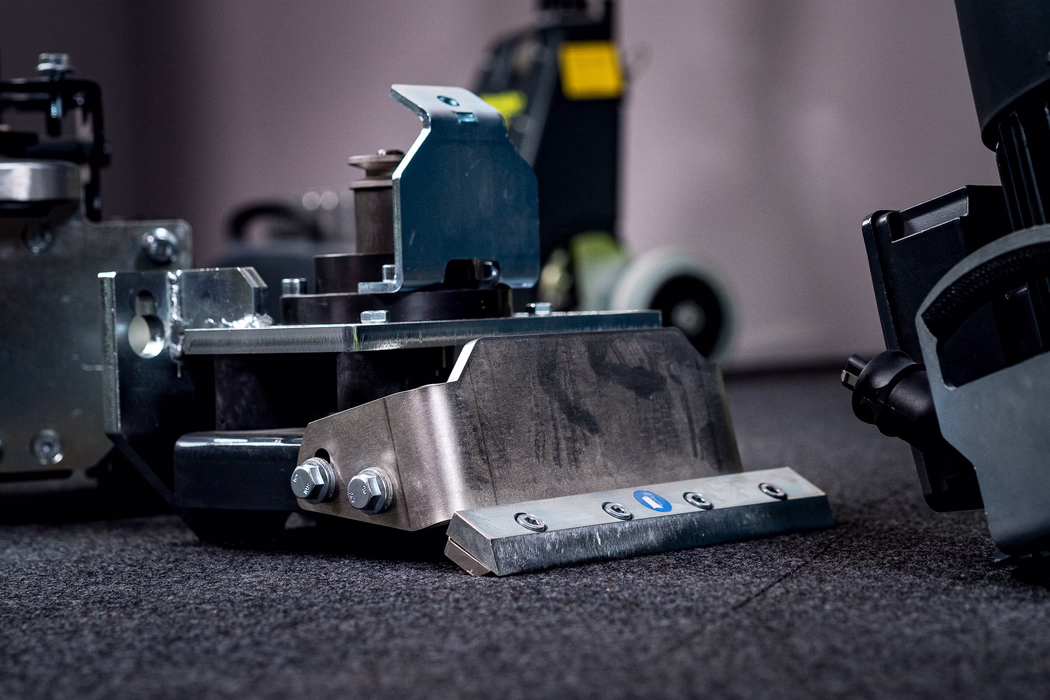 Close-up of a heavy metal industrial component placed on a dark, textured carpet. In the foreground a sturdy stainless steel bracket with two large hex bolts is attached to a beveled metal scraping blade fixed by four countersunk screws; a small blue circular sticker is visible on the blade. The metal surfaces show fine machining marks, slight wear and faint oil or grease smudges. Behind the main part, other machine elements and a caster wheel are visible but blurred by a shallow depth of field. Cool, directional lighting emphasizes the metallic textures, edges and reflections, and the low-angle composition highlights the component's solid, utilitarian construction.
